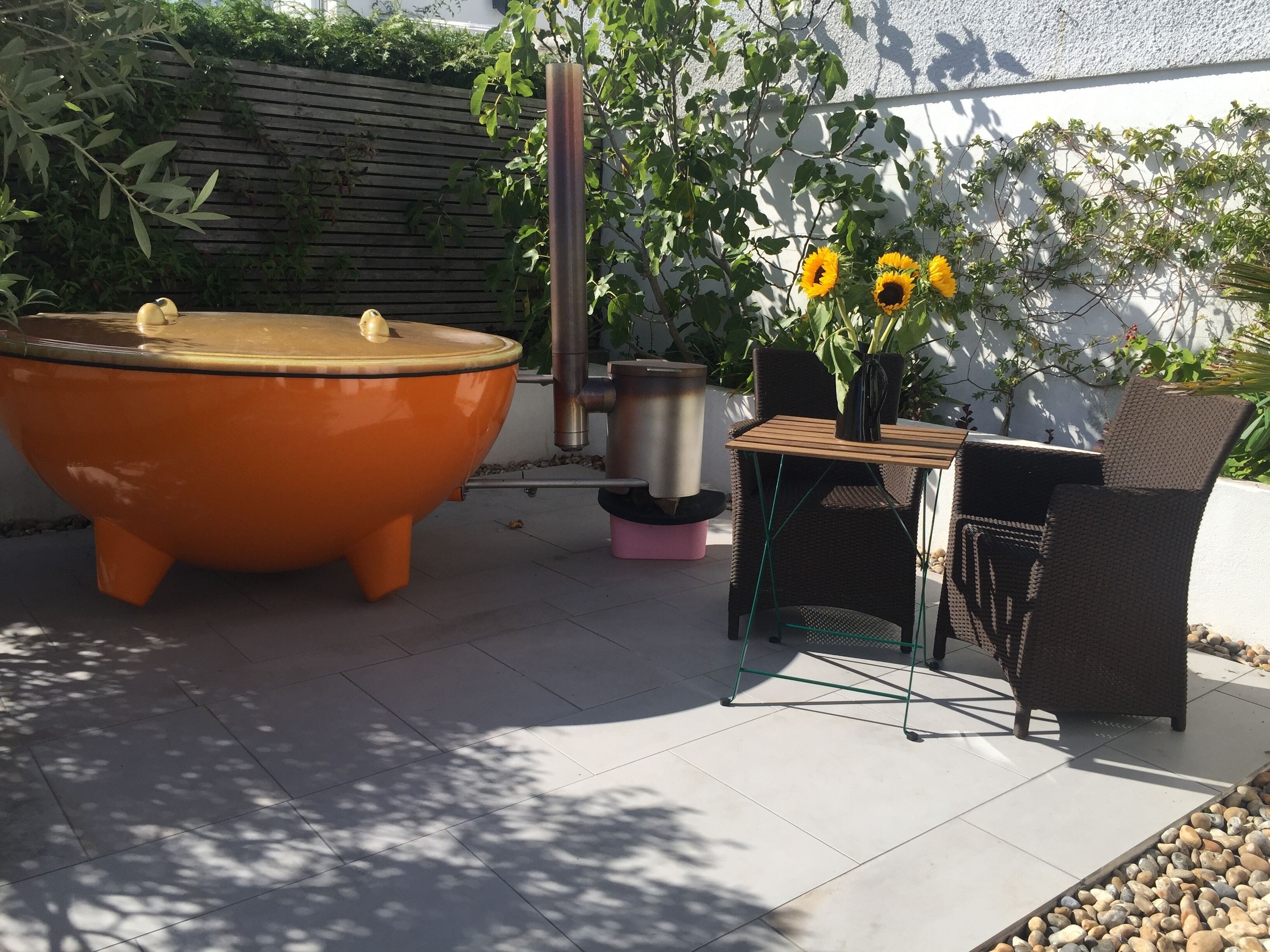 En udendørs terrasse med en stor terracotta-potte, et bord med stole og en potteplante med solsikker.