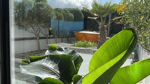 A view through a window of a courtyard with large green plants, a blue fence, and a clear sky. - The Seahorse Torbay - Seaside hideaway for two with hot tub and movie room (Torquay)
