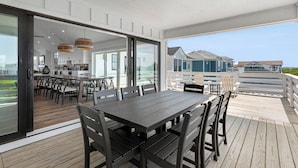 Een moderne eethoek buiten met een grote tafel en stoelen, een keuken op de achtergrond en uitzicht op omliggende huizen.
