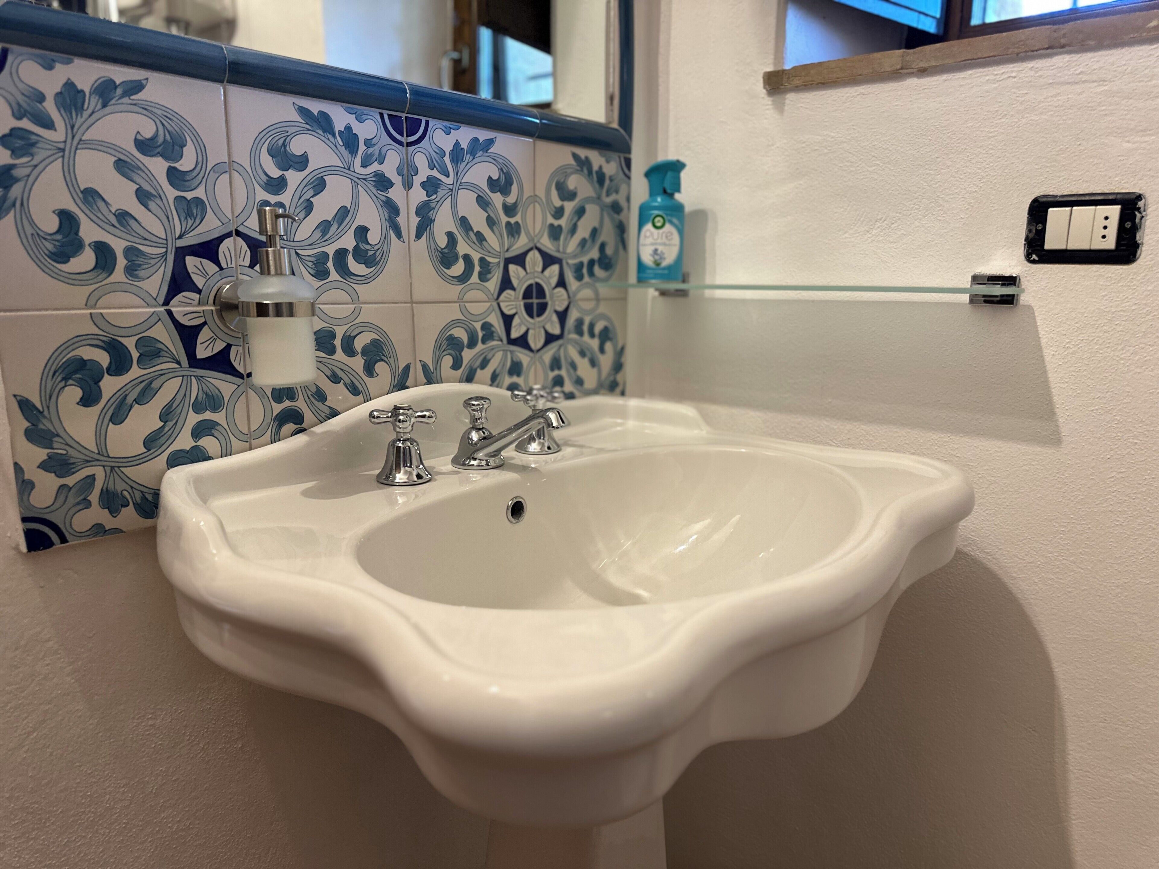 Une salle de bain avec un lavabo blanc, des carreaux à motifs bleus et blancs, un distributeur de savon et une étagère en verre.