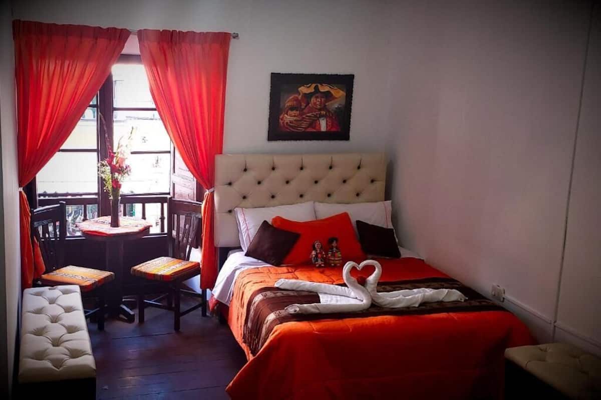 A hotel room with a bed, a small table with chairs, a bench, and a window with curtains.