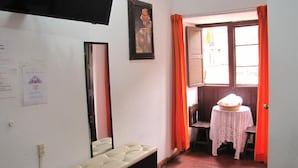 A room with a bed, a table with a basket, a chair, a window with curtains, and a wall-mounted TV. - Imperial Golden House Inn (Cusco)