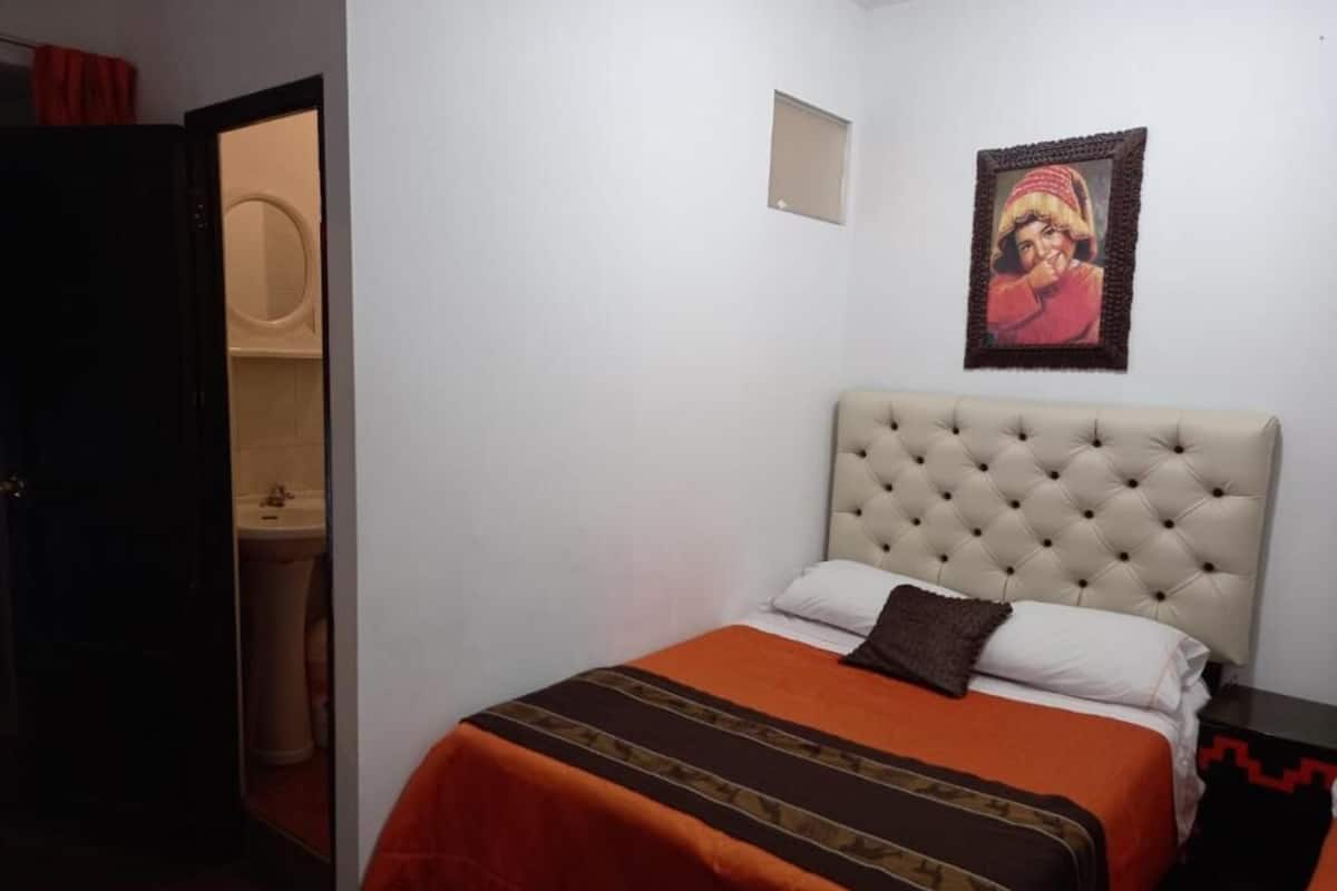 A bedroom with a tufted headboard, an orange and brown striped bedspread, a small framed picture on the wall, and a partially open door leading to a bathroom.