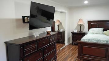 Habitación de hotel con un televisor de pantalla plana grande montado en la pared, un aparador de madera con varios cajones, dos mesitas de noche con lámparas y una cama con ropa de cama estampada.