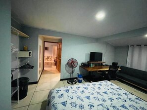 Ein Hotelzimmer mit Bett, Schreibtisch, Stuhl, Fernseher, Ventilator und einer Tür zu einem anderen Raum.