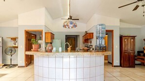 A kitchen with a central island, wooden cabinets, a fan, and a water dispenser. - Casa Neju Diri - surf digital nomad hideaway (Nosara)