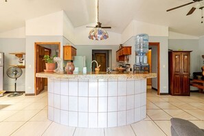 A kitchen with a central island, wooden cabinets, a fan, and a water dispenser. - Casa Neju Diri - surf digital nomad hideaway (Nosara)