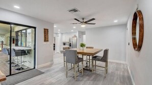 Uma zona de refeições moderna com mesa e cadeiras de madeira, ventoinha no teto e vista para a cozinha.