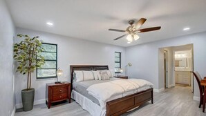 Quarto com cama, mesas de cabeceira, ventilador de teto, planta em vaso e vista para o banheiro.