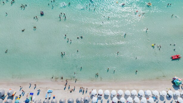 En vy över en strand från ovan, där man ser folk som badar och solar, och rader av parasoller och solstolar längs stranden.