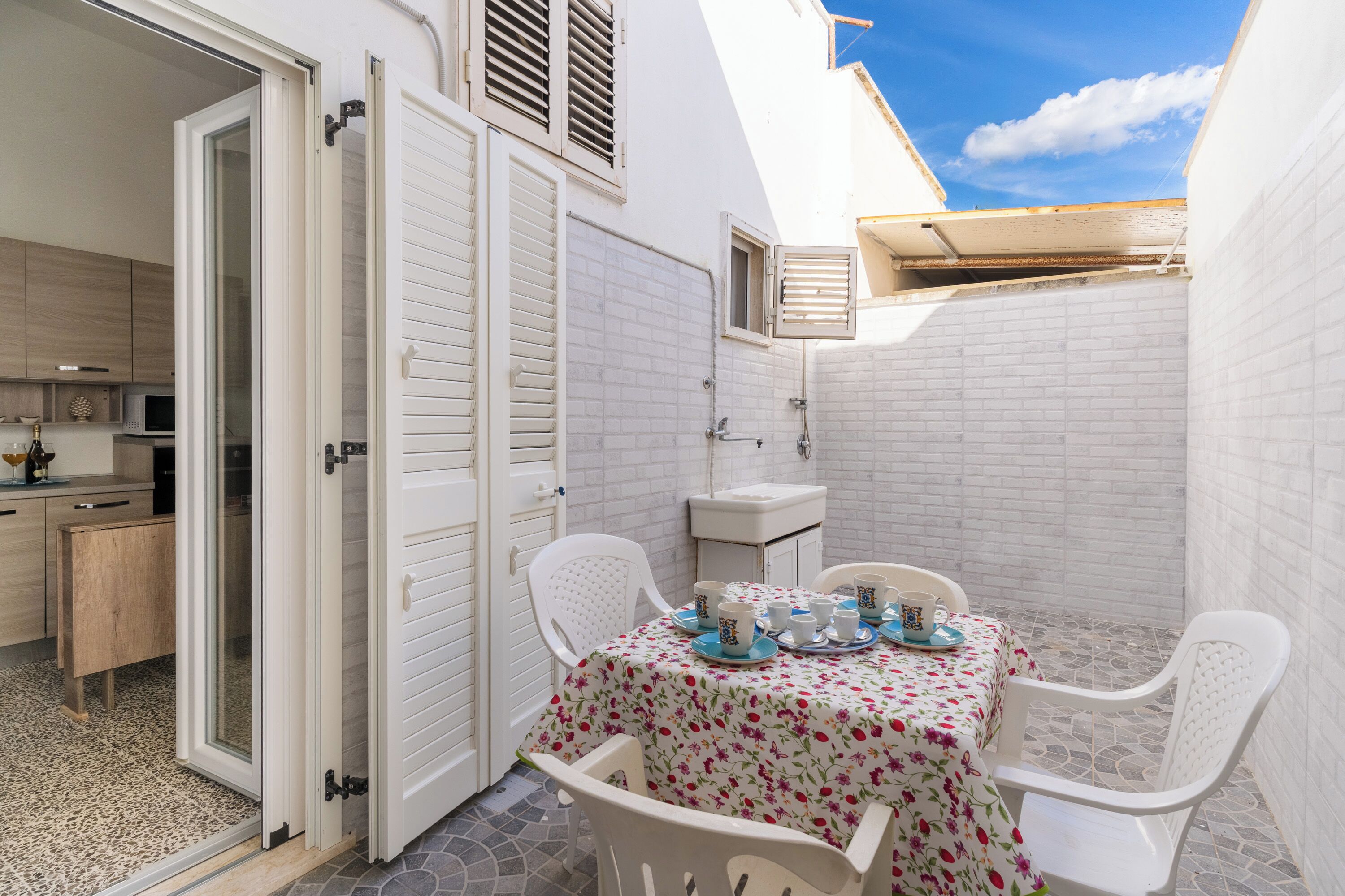 A small, enclosed outdoor dining area with a table set for four, surrounded by white chairs, a white sink, and a small window.