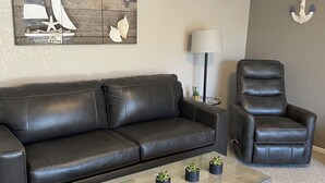 A living room with a black leather sofa, a glass coffee table with potted plants, a black leather armchair, a lamp, and a nautical-themed wall art. - Location Location! Cute cozy condo! Close to everything! (Osage Beach)