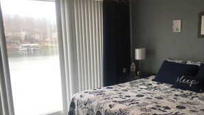 A bedroom with a large bed, a nightstand, a lamp, a window with blinds, and a view of a lake. - Location Location! Cute cozy condo! Close to everything! (Osage Beach)