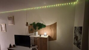 A room with a flat-screen TV on a wooden stand, a potted plant, and a wall-mounted shelf with a lamp. - Magnifique Studio Au Petit Nid Douillet Face Mer est Port (Narbonne)