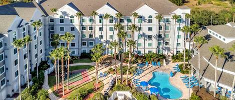 A multi-story hotel with a pool area, palm trees, and a tennis court.