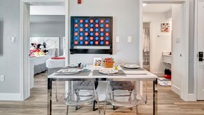 Un coin repas avec une table dressée pour quatre personnes, des chaises transparentes, une grande œuvre d’art abstrait accrochée au mur et, au fond, un lit avec des jouets.