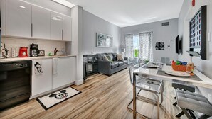 Une cuisine moderne avec des meubles blancs, un coin repas doté d’une table en verre et un canapé gris dans le salon.