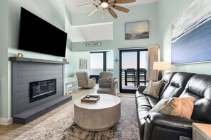A living room with a fireplace, a flat-screen TV, a leather sofa, and a view of the outdoors. - 🏝️Sound-Front | Two Balconies | Steps to the Beach | Palm Beach Club #206🏝️ (Pensacola Beach)