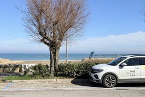 Een witte auto geparkeerd bij een boom, met een strand en zee op de achtergrond.
