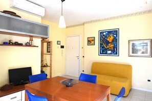 Een kamer met een houten eettafel, blauwe stoelen, een gele bank, een televisie en kunstwerken aan de muren.