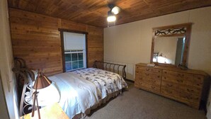 Een slaapkamer met een houten plafond, een bed met een bloemenbedekking, een kaptafel met spiegel en een raam met zonwering.