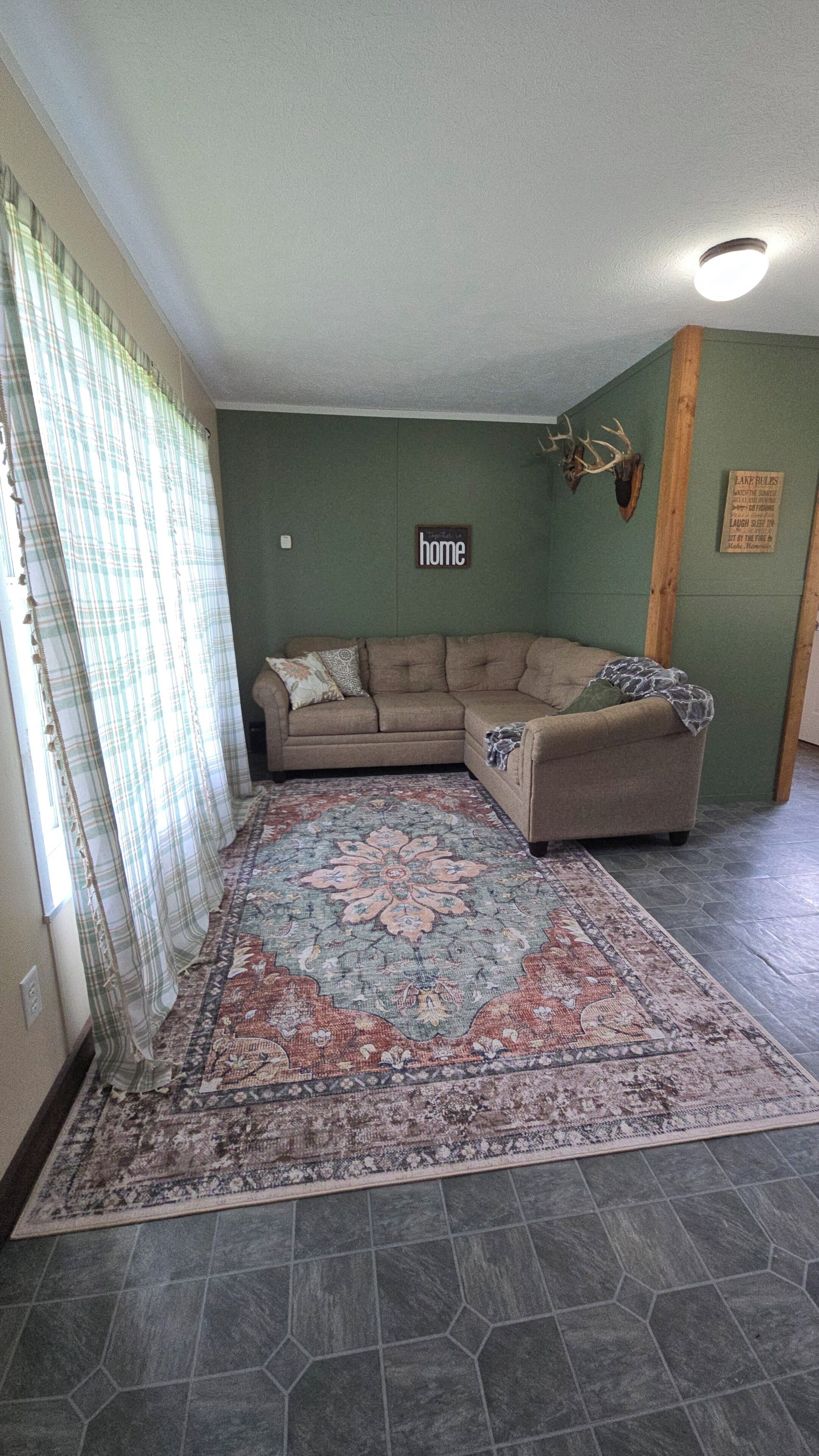 Un salon avec un canapé d’angle, un tapis à motifs, une tête de cerf accrochée au mur et une pancarte portant l’inscription « chez soi ».