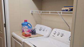 Un espace buanderie comprenant un lave-linge et un sèche-linge à chargement frontal, une étagère avec une boîte étiquetée « Très froid », et une bouteille de lessive Downy.