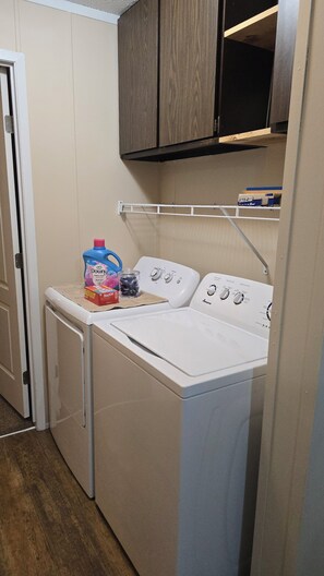 Een wasruimte met een voorlader wasmachine en droger, een plank met een doos waarop 'Koud Koud' staat, en een fles Downy wasverzachter.