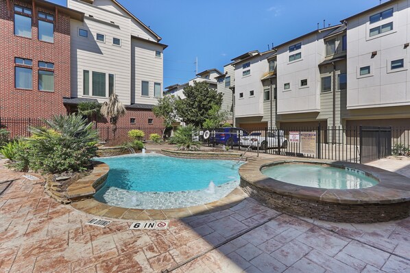 Un edificio residencial con piscina y jacuzzi, rodeado por un muro de ladrillo y vegetación.