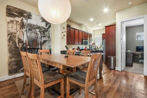 Un comedor con una mesa y sillas de madera, un cuadro grande en la pared y una cocina al fondo.