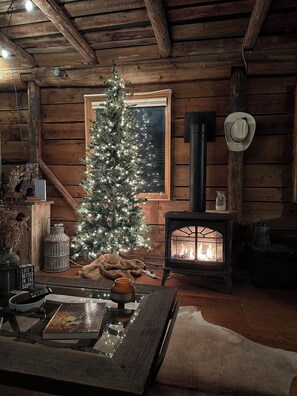 L’intérieur chaleureux d’un chalet avec un mur en rondins de bois, un sapin de Noël illuminé, un poêle à bois et une table à manger rustique.