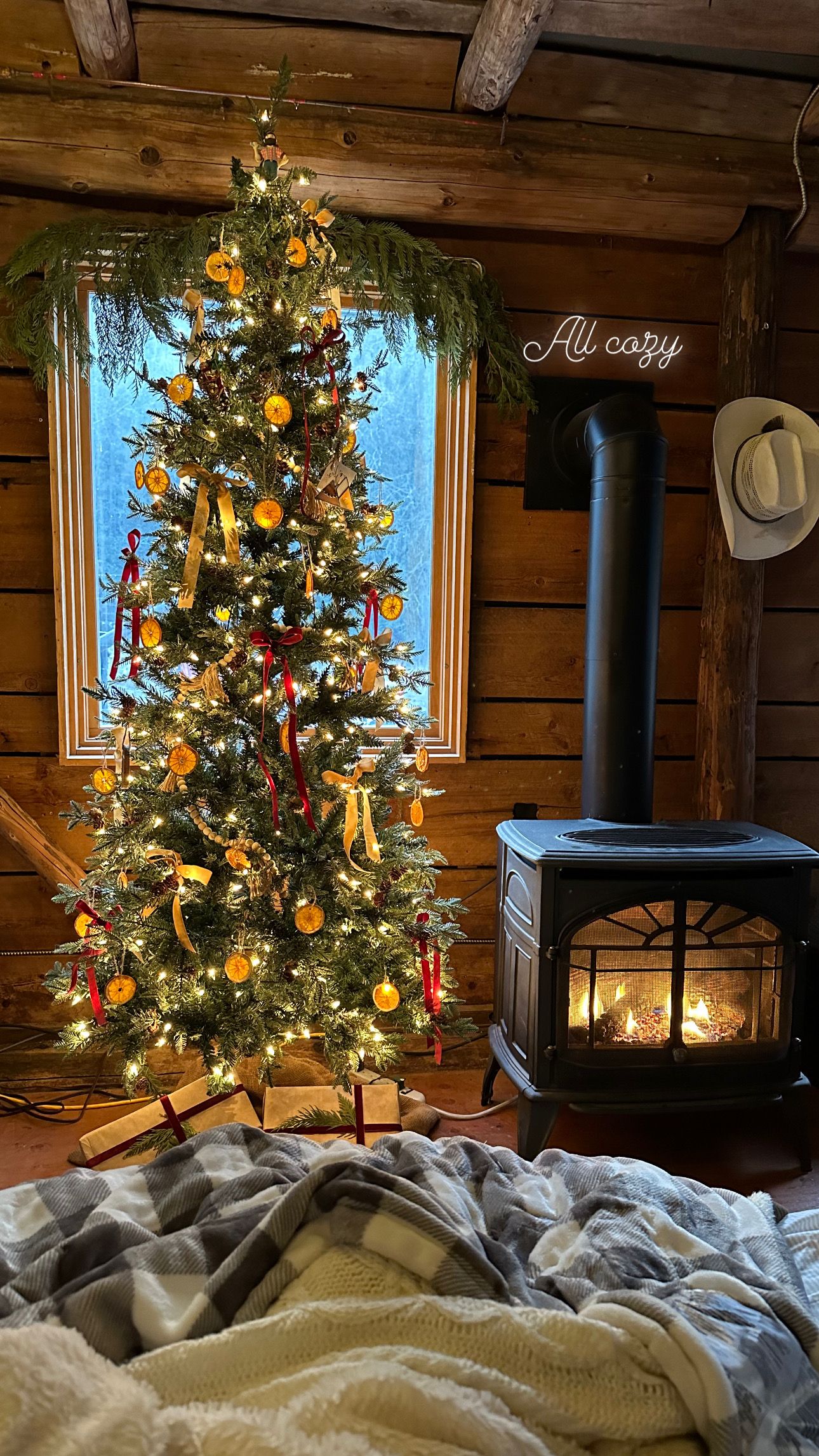 L’intérieur chaleureux d’un chalet avec un sapin de Noël décoré, un poêle à bois et une fenêtre offrant une belle vue.