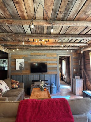 Un intérieur rustique avec des poutres apparentes, un téléviseur fixé au mur, un canapé agrémenté de coussins décoratifs et un plaid rouge posé sur le sol.