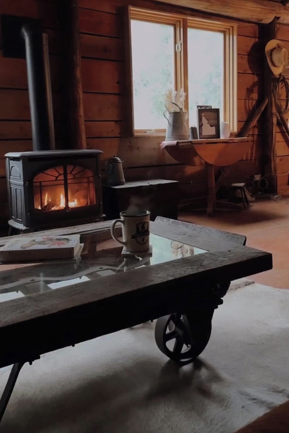 Interior rústico de una cabaña con estufa de leña, una mesa con una taza y un ventanal con vistas al exterior.