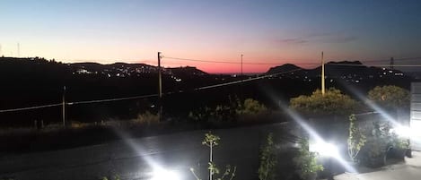 A rooftop with potted plants and a view of a sunset over a hilly landscape.