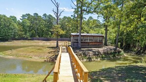 A wooden bridge leading to a cabin with large windows, surrounded by a forest and a pond.