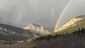 A mountain landscape with a rainbow, a clear sky, and a few buildings in the foreground.