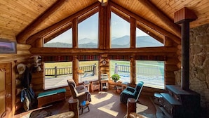 A log cabin interior with a high ceiling, large windows, a fireplace, and a view of the outdoors.