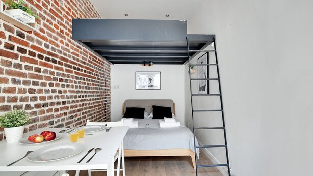 A compact bedroom with a brick wall, a bed with a headboard, a ladder, a small table with breakfast items, and a potted plant. - APPARTEMENT PARIS 10eme CANAL ST MARTIN (Paris)