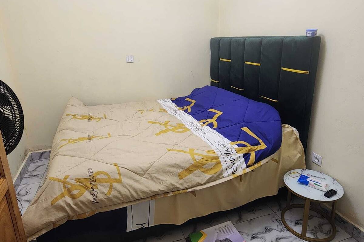 Ein kleines Zimmer mit einem Bett, einem Ventilator, einem kleinen Tisch mit Gegenständen und einer Steckdose.