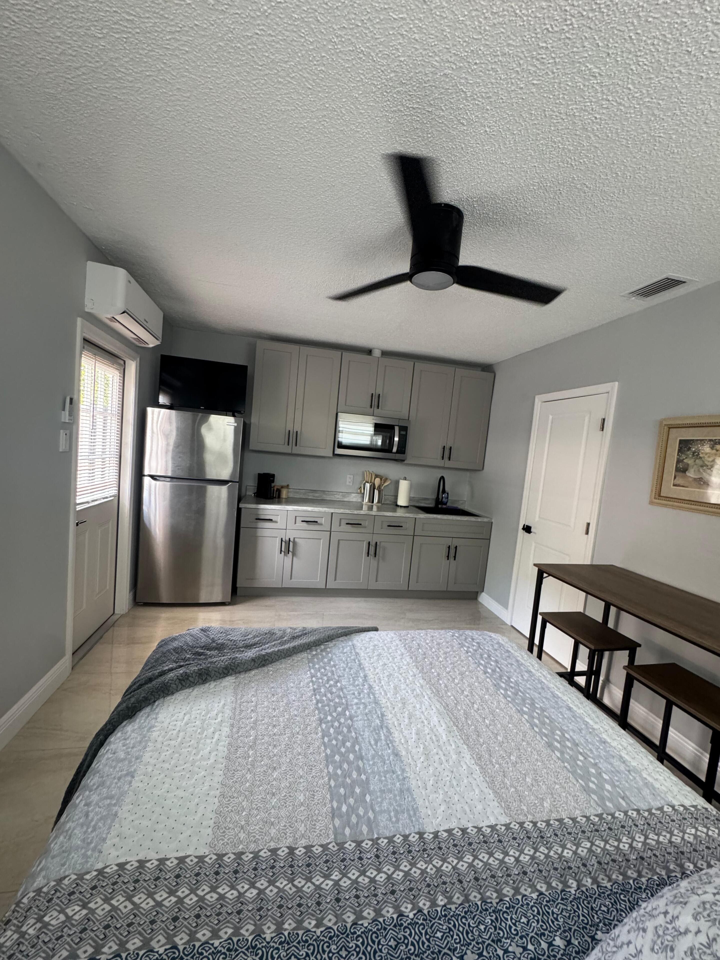 Un dormitorio con cama, ventilador de techo, kitchenette con refrigerador y microondas, y una puerta que da a otra habitación.