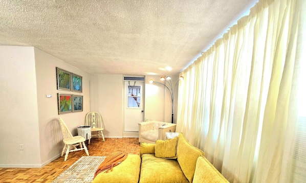 A living room with a yellow sofa, a white chair, a small table, and a lamp. There are framed pictures on the wall and a door with a window. - Cozy haven (Toronto)