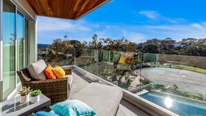 A balcony with a wicker sofa, cushions, a small table with plants, and a view of a parking area and trees.