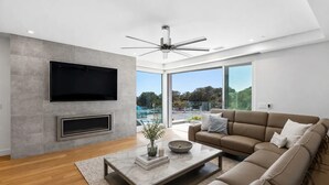 A modern living room with a flat-screen TV, a fireplace, a ceiling fan, a large window with a view of a pool and trees, and a beige sofa.