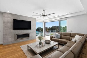 A modern living room with a flat-screen TV, a fireplace, a ceiling fan, a large window with a view of a pool and trees, and a beige sofa.