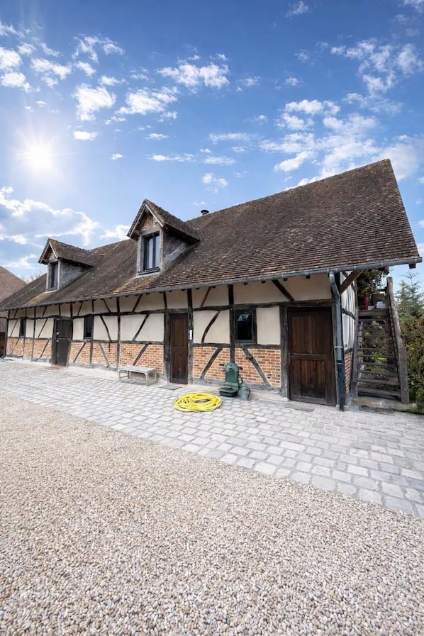 Ein traditionelles Fachwerkhäuschen mit einem Kieshof, Holzbanken und einem gelben Gartenschlauch auf dem Boden.