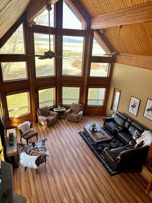 A spacious living room with large windows, a fireplace, and a high ceiling.