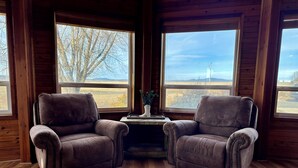 A room with two brown leather armchairs facing a wooden table with a potted plant. Large windows offer views of a landscape with trees and a distant structure.