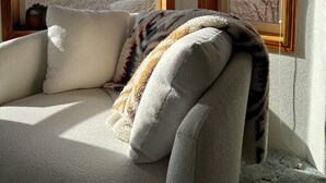 A room with a sofa, a window, and a view of a field and mountains.