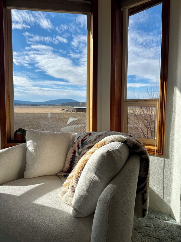 A room with a sofa, a window, and a view of a field and mountains.